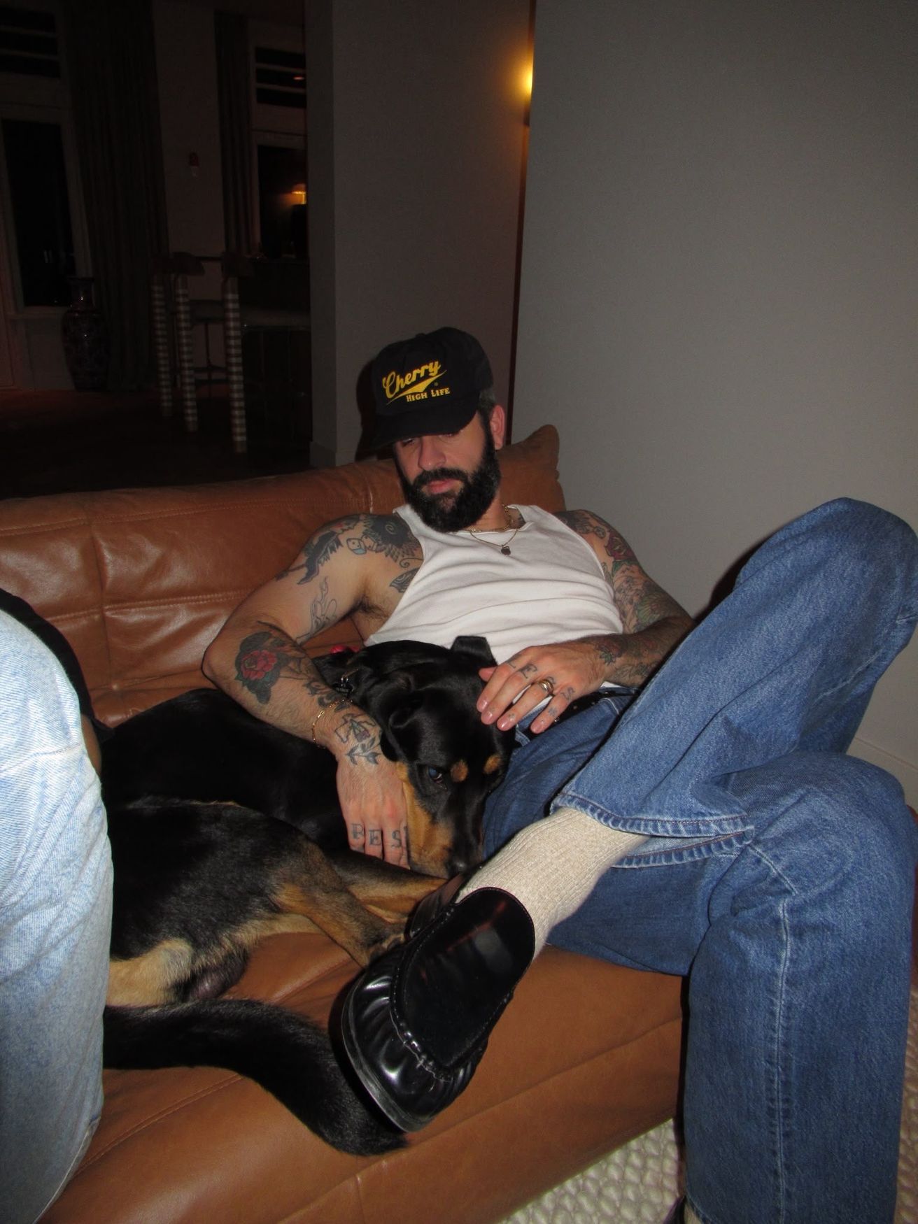 Joey on a leather sofa with his dog, in jeans and a white tank top